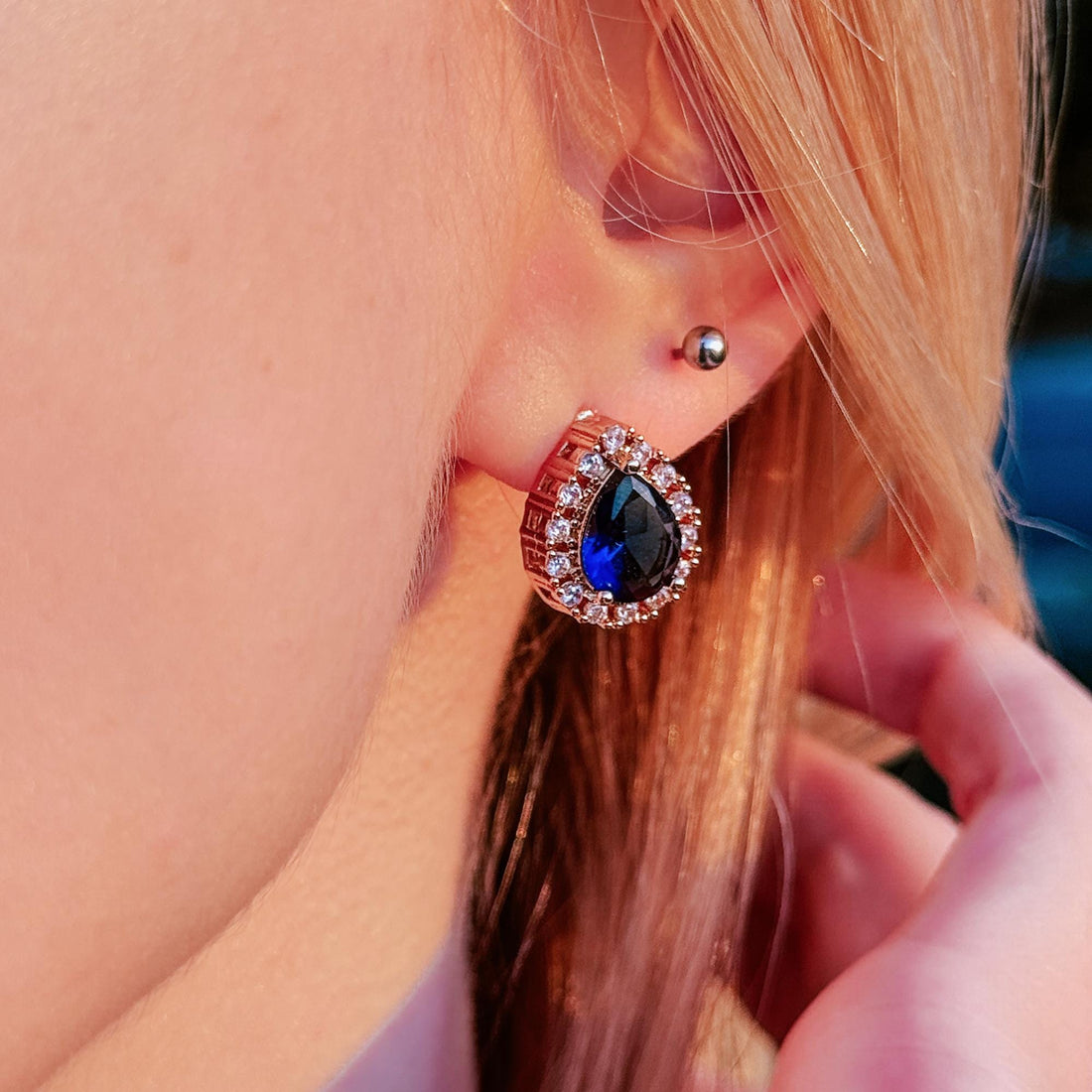 May include: Rose gold teardrop earrings with a dark blue center stone and a halo of small, clear stones. The earrings are set in a square frame and are worn on a person's ear.