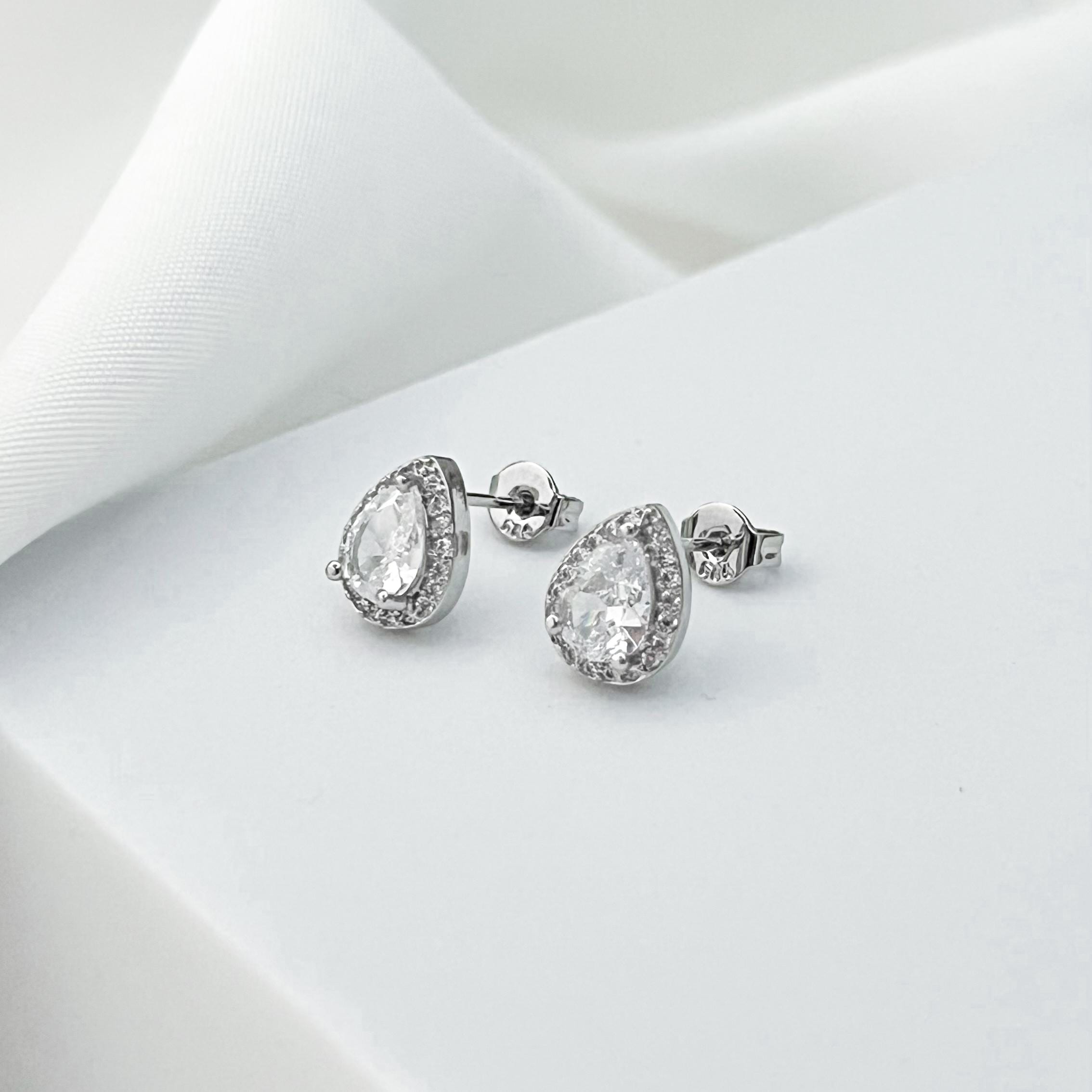 May include: A pair of silver stud earrings with a pear-shaped, clear gemstone center, surrounded by a halo of smaller, clear stones. The earrings are displayed on a white surface, with a soft, white fabric in the background.