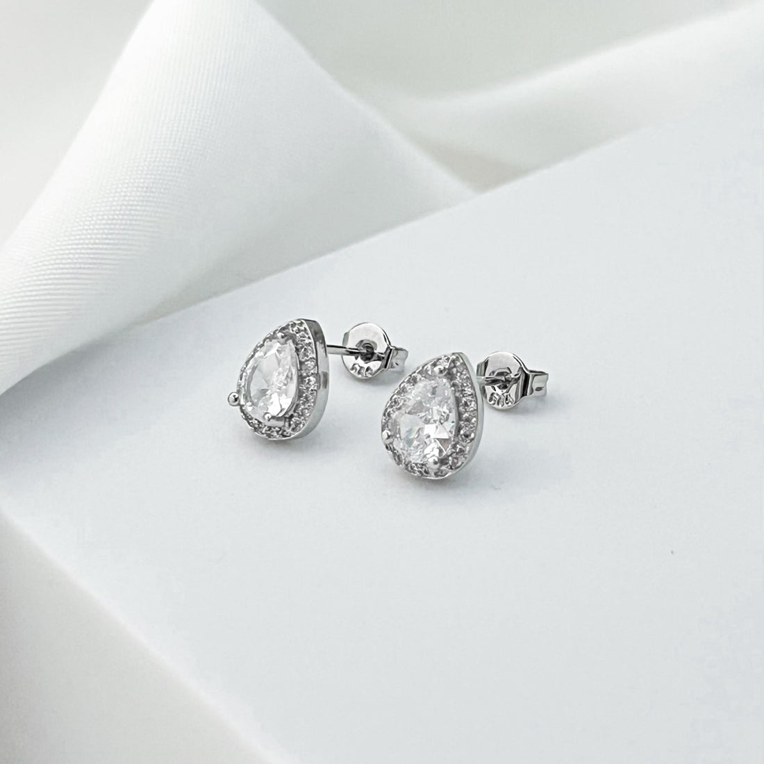 May include: A pair of silver stud earrings with a pear-shaped, clear gemstone center, surrounded by a halo of smaller, clear stones. The earrings are displayed on a white surface, with a soft, white fabric in the background.